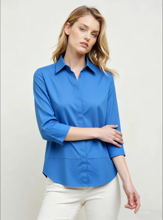 Woman wearing a bright blue collared shirt with 3/4 sleeves and white pants, standing with arms crossed.