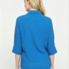 Back view of a woman wearing a bright blue collared shirt with 3/4 sleeves and white pants