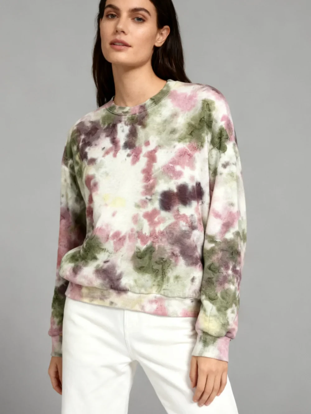 Woman wearing a multicolor (pink, green, purple) tie - dye sweatshirt with white pants against a gray background.