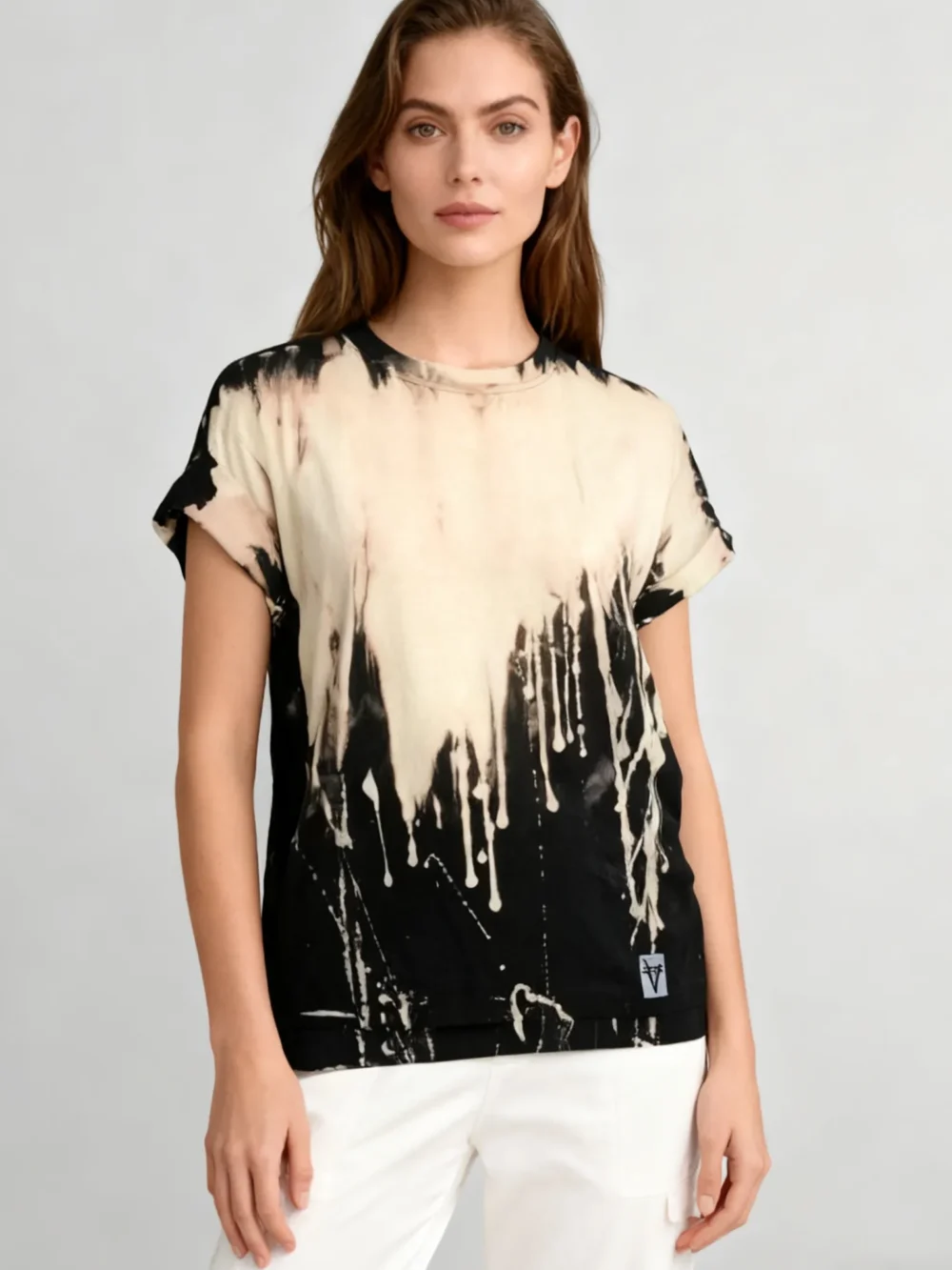 Woman wearing a black and beige drip - print tie - dye short - sleeve T - shirt with white pants against a light gray background.