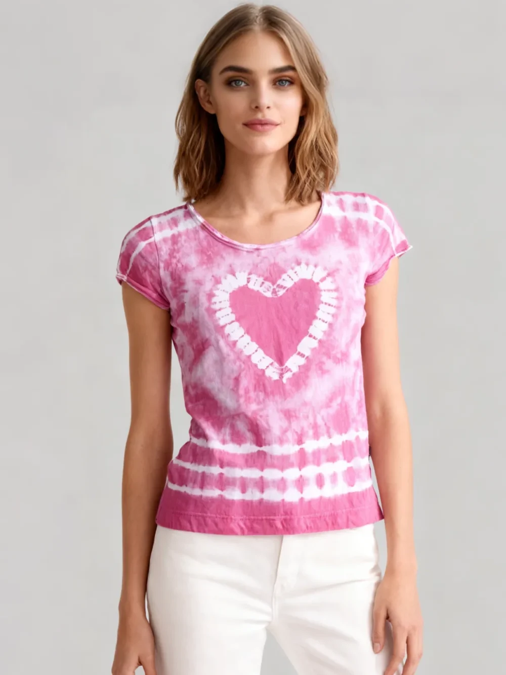 Woman wearing a pink tie - dye heart print short sleeve t - shirt with white pants against a light gray background.