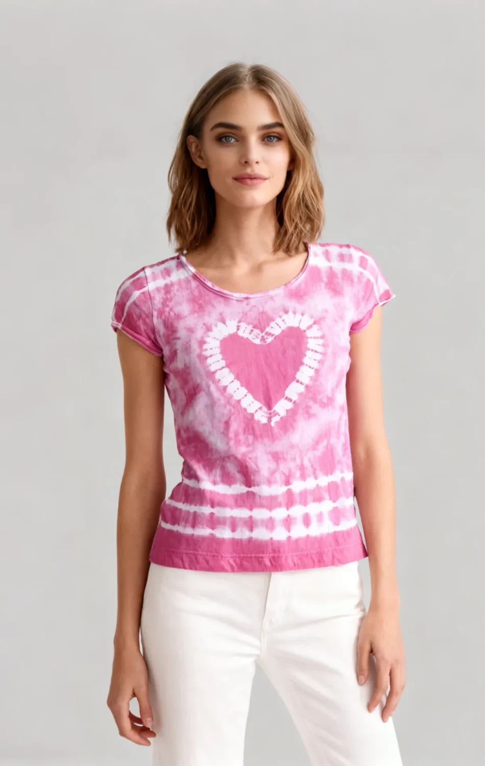 Woman wearing a pink tie - dye heart print short sleeve t - shirt with white pants against a light gray background.