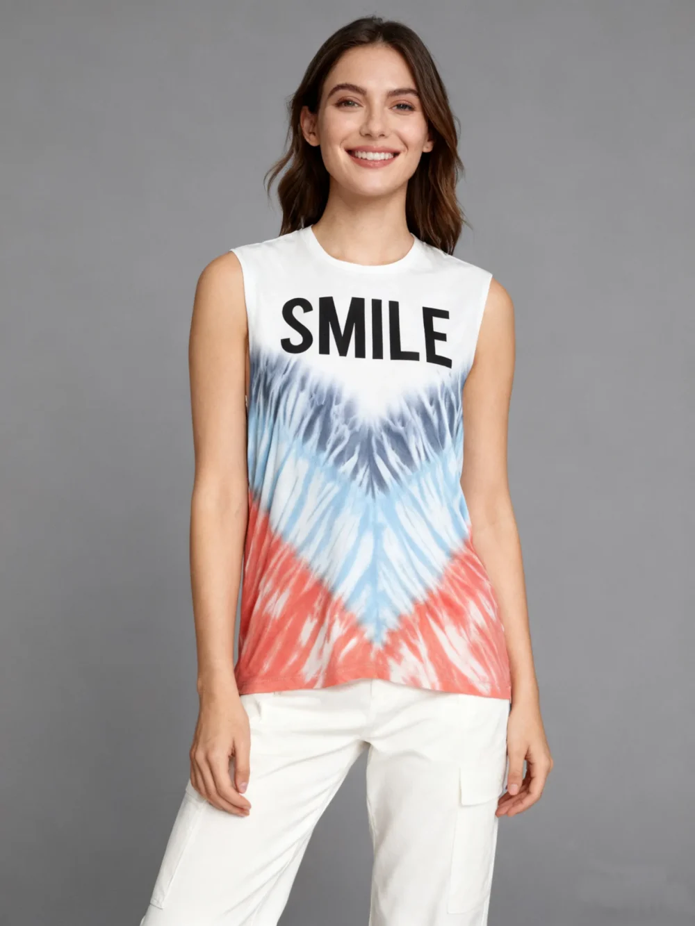 Woman wearing a “SMILE” graphic tie - dye sleeveless tank top with white pants against a gray background.