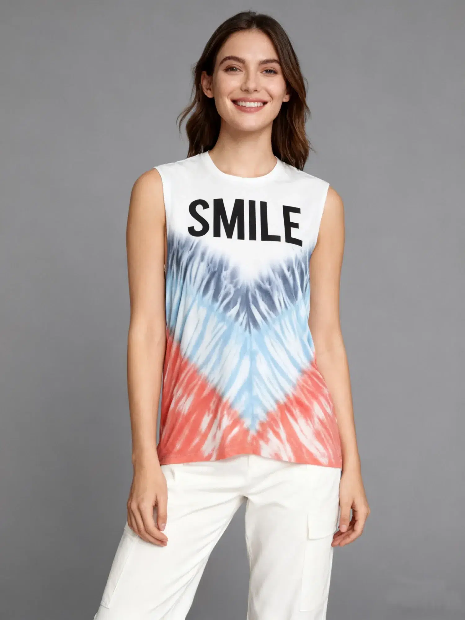 Woman wearing a “SMILE” graphic tie - dye sleeveless tank top with white pants against a gray background. Woman wearing a “SMILE” graphic tie - dye sleeveless tank top with white pants against a gray background.