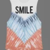 Woman wearing a “SMILE” graphic tie - dye sleeveless tank top with white pants against a gray background. Woman wearing a “SMILE” graphic tie - dye sleeveless tank top with white pants against a gray background.
