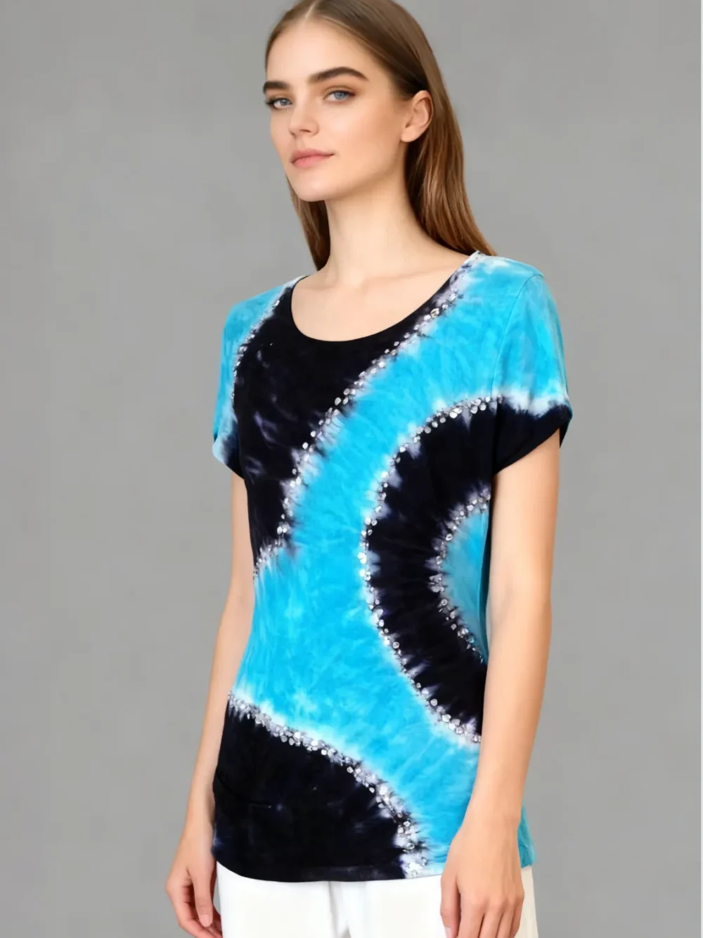 Woman wearing a blue and black tie - dye short sleeve top with rhinestone embellishments, paired with white pants against a gray background.