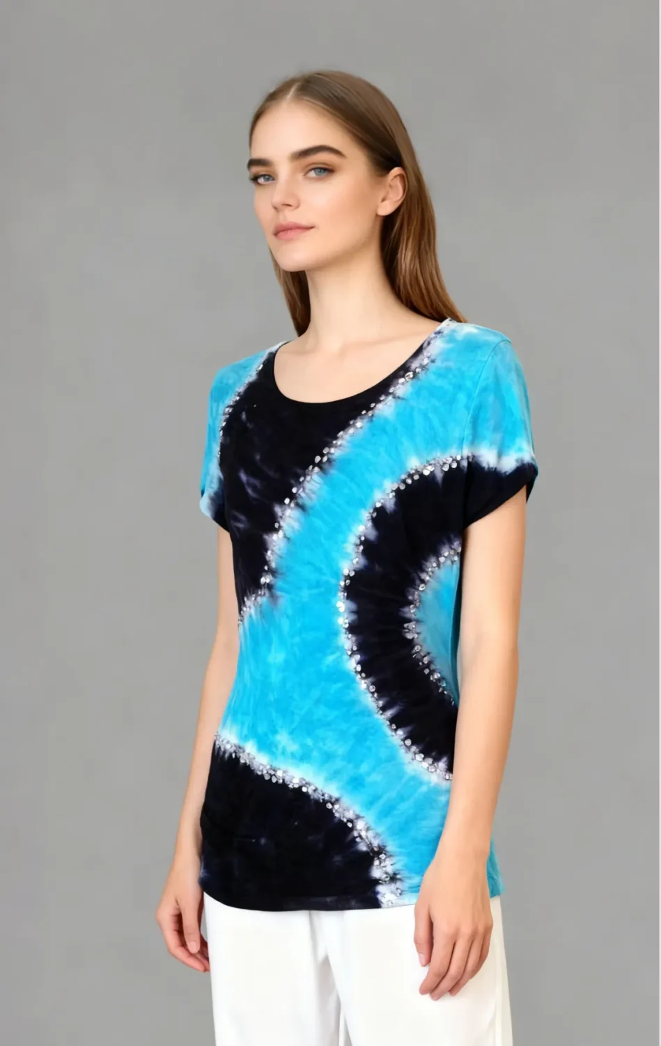 Woman wearing a blue and black tie - dye short sleeve top with rhinestone embellishments, paired with white pants against a gray background.