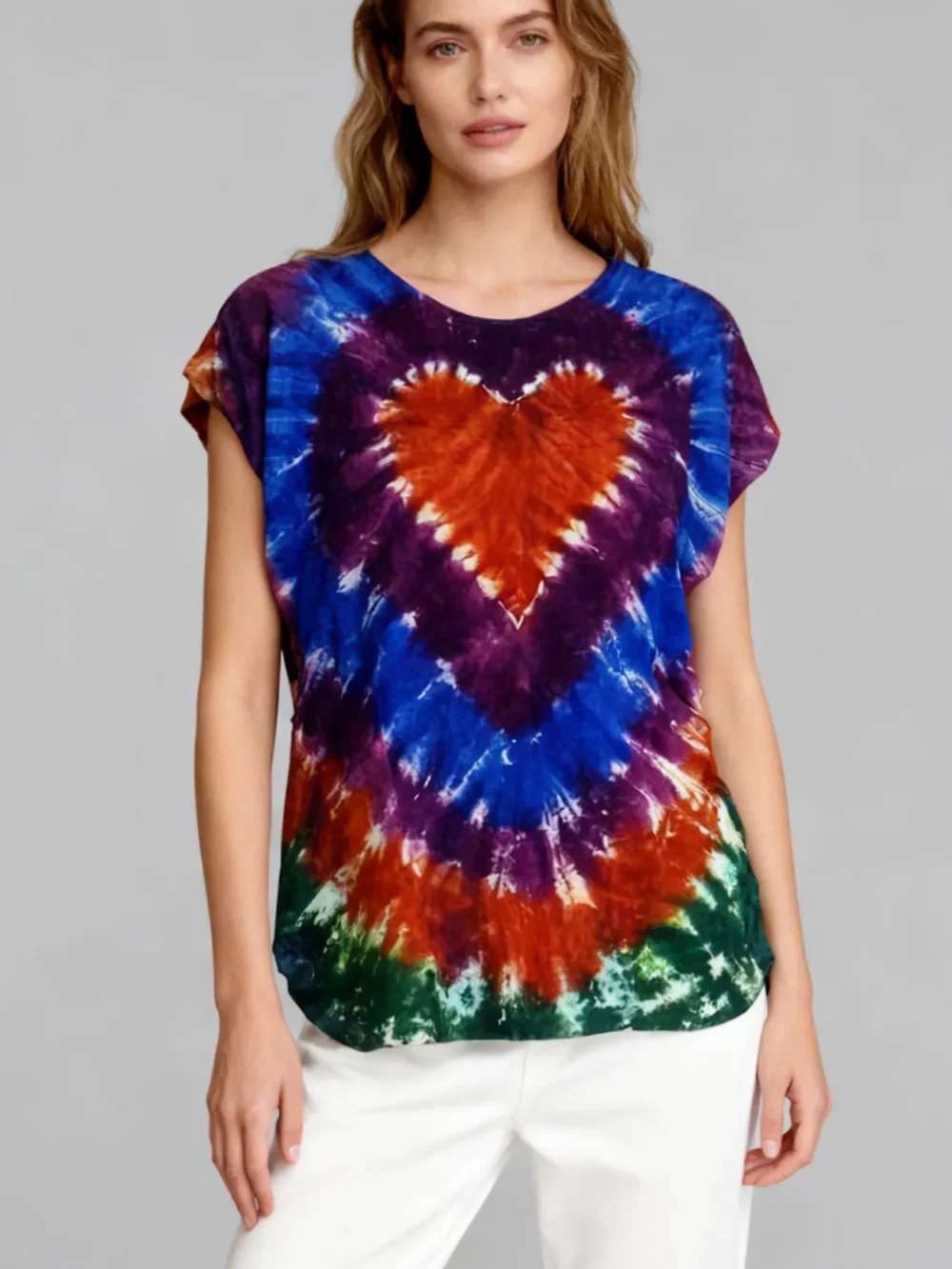 Woman wearing a multicolor heart tie - dye short sleeve top with white pants against a light gray background.