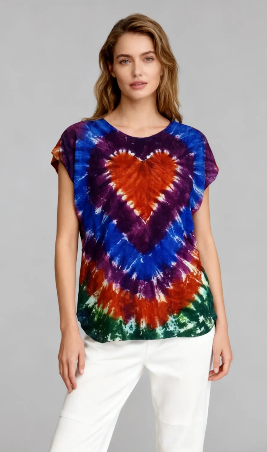 Woman wearing a multicolor heart tie - dye short sleeve top with white pants against a light gray background.