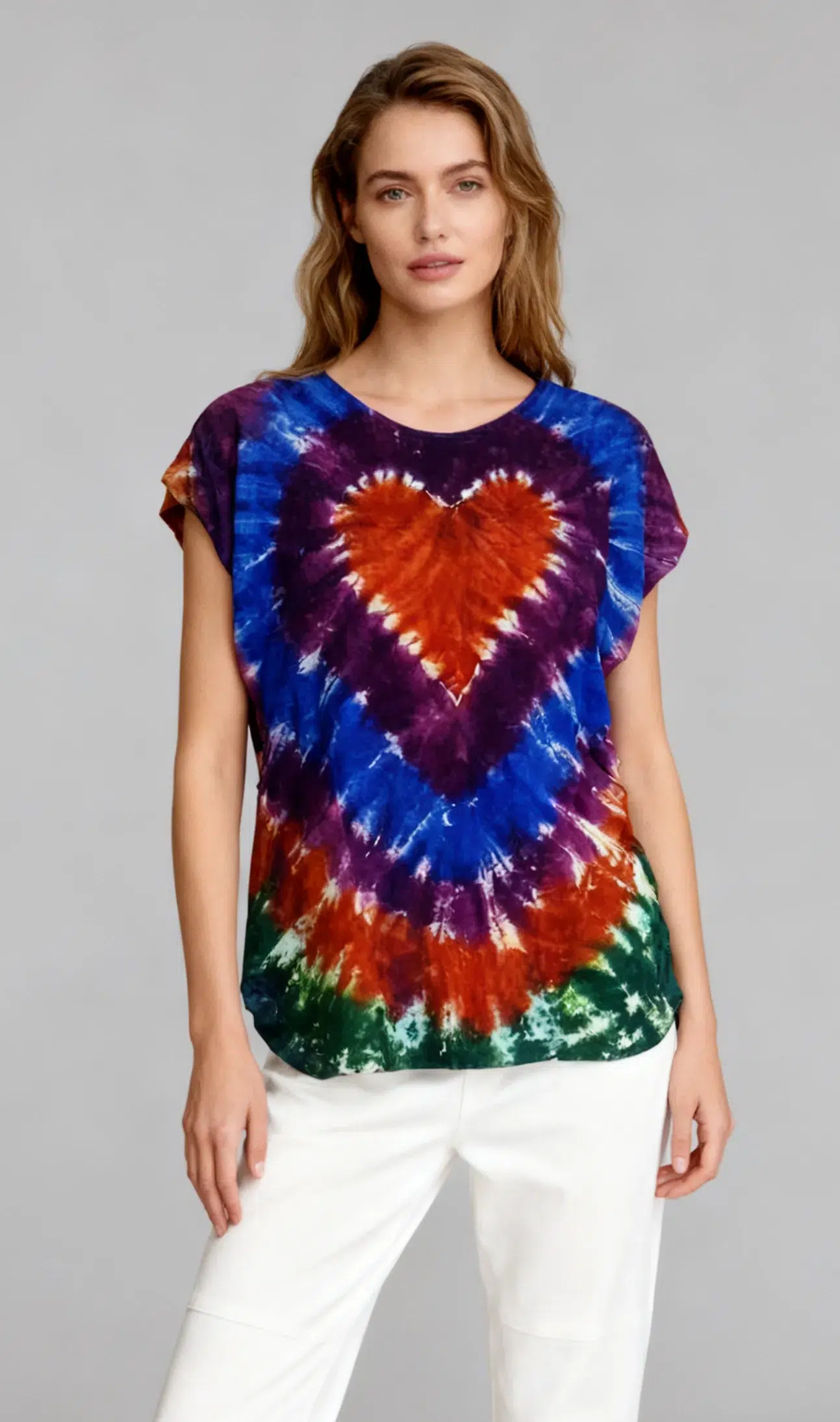 Woman wearing a multicolor heart tie - dye short sleeve top with white pants against a light gray background.