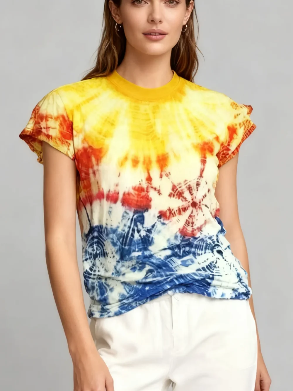 Woman wearing a rainbow burst tie - dye short sleeve top with white pants against a light gray background.