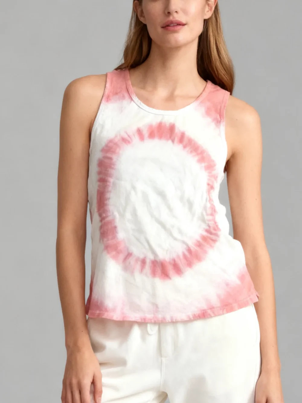 Woman wearing a pink and white spiral tie - dye sleeveless tank top with white pants against a light gray