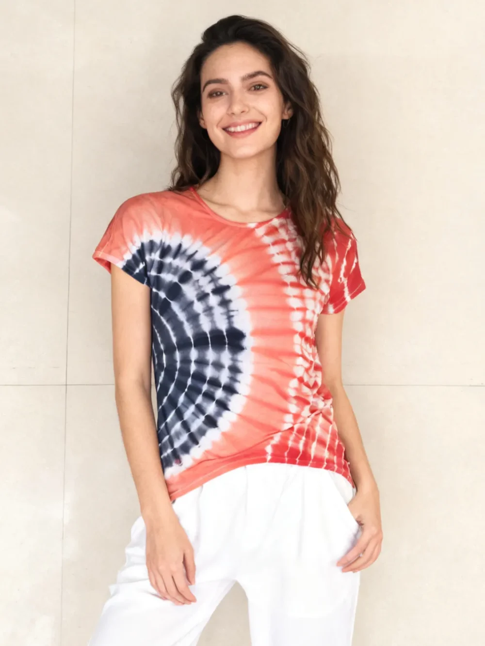 Woman wearing a red and navy spiral tie - dye short sleeve top with white pants against a light beige tiled wall.