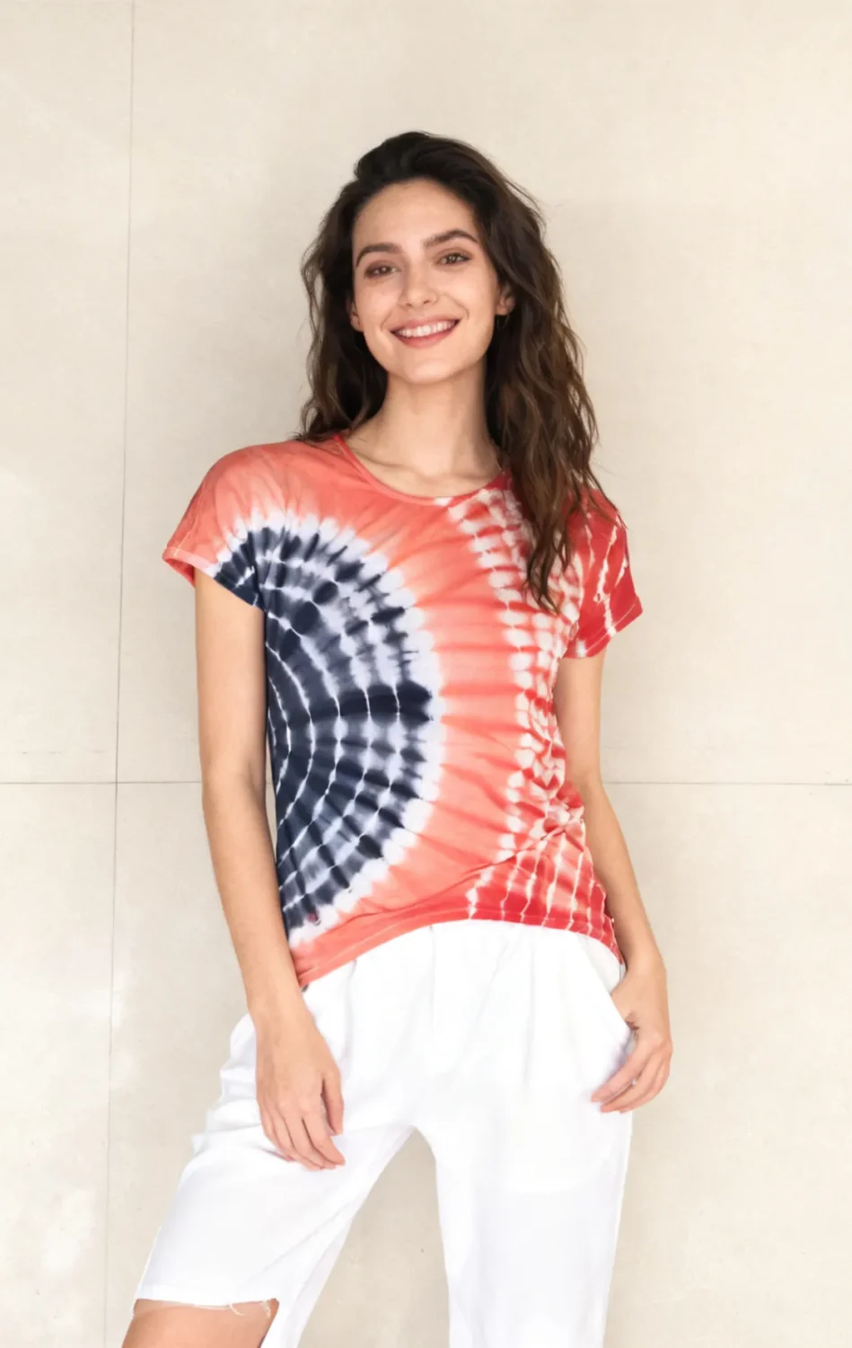 Woman wearing a red and navy spiral tie - dye short sleeve top with white pants against a light beige tiled wall.