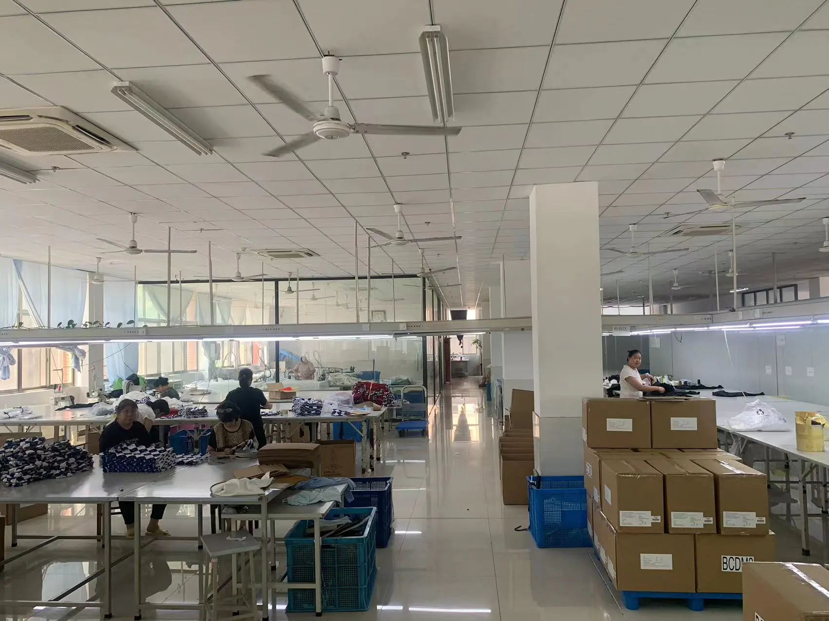 Garment factory inspection department with workers, fabrics & packaged goods, suitable for quality inspection & finished product handling.